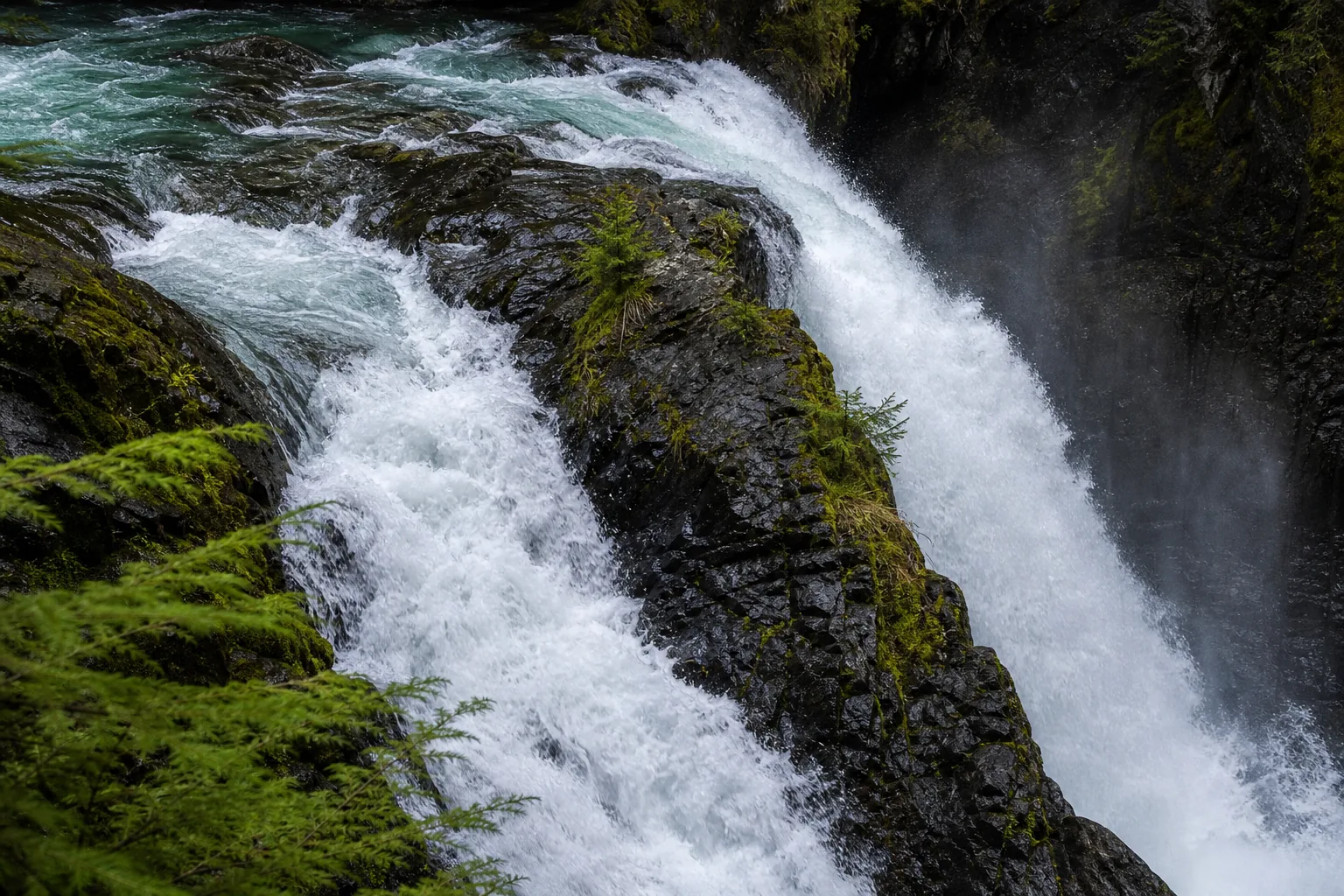 Detail of water hitting dark basalt cliffs in the Nooksack Falls gorge with moss along the rim